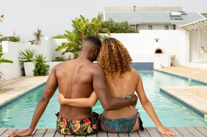 Rear view of young African-american couple sitting together near swimming pool at the backyard of home. Summer fun at home by the swimming pool