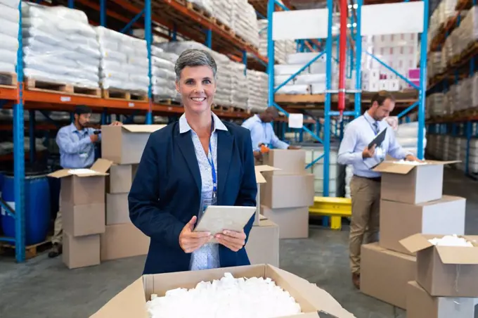 Front view of happy mature Caucasian female manager with digital tablet looking at camera in warehouse. Diverse mature warehouse worker unpacking cardboard boxes. This is a freight transportation and distribution warehouse. Industrial and industrial workers concept