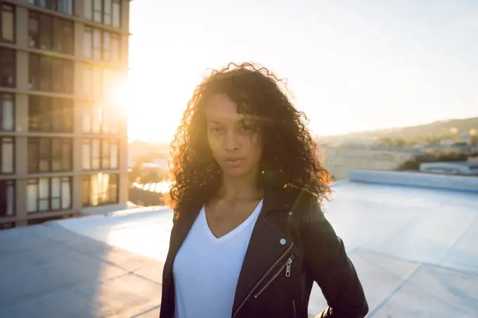 Front view of a young African-American woman wearing a leather jacket looking intently at the camera while standing on a rooftop with a view of a building and the sunset