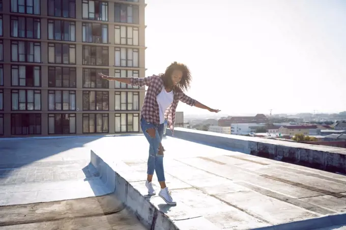 Front view of a young African-American woman wearing a plaid jacket with hands stretched while walking on a rooftop with a view of a building and sunlight