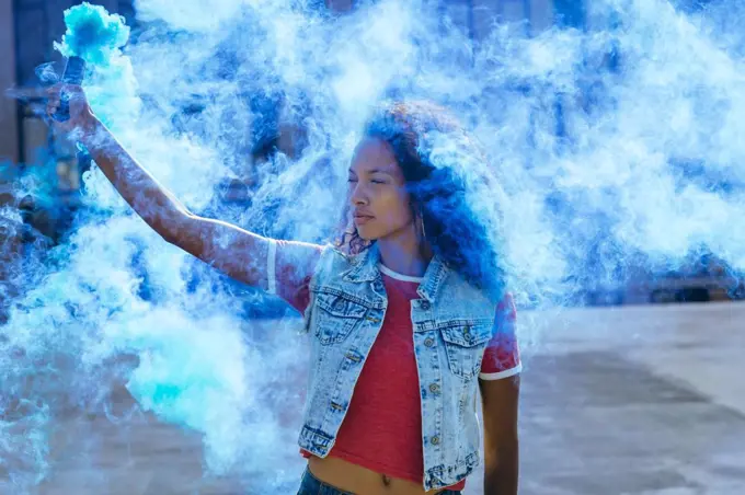 Front view of a young African-American woman wearing a denim vest holding a smoke maker producing blue  smoke