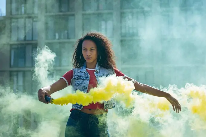 Front view of a young African-American woman wearing a denim vest holding a smoke maker producing yellow smoke  on a rooftop with a view of a building and sunlight