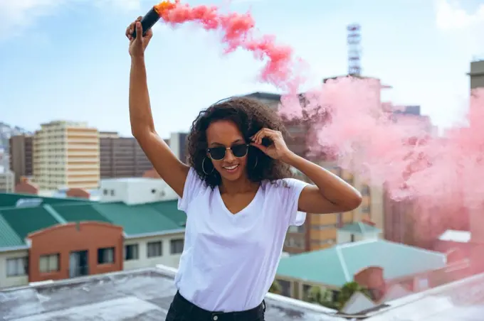 Front view of a young African-American woman wearing a white shirt and eyeglass smiling while holding a smoke maker producing red smoke on a rooftop with a view of buildings. Bright modern gym with fit healthy people working out and training