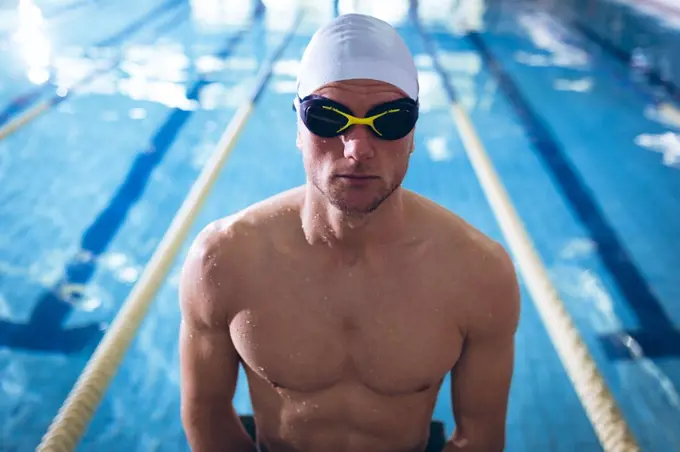Front view of a male Caucasian swimmer wearing a white swimming cap and goggles standing by an olympic sized pool inside a stadium