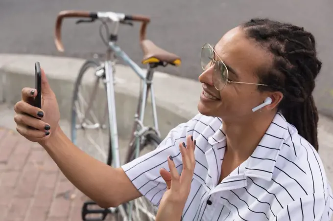 Side view close up of a mixed race man with long dreadlocks out and about in the city on a sunny day, sitting in the street wearing headphones, using a smartphone and waving his hand, with his bicycle standing next to him. 