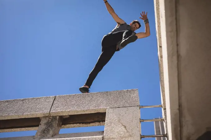 Front low angle view of a Caucasian man practicing parkour by the building in a city on a sunny day, jumping on the rooftop.