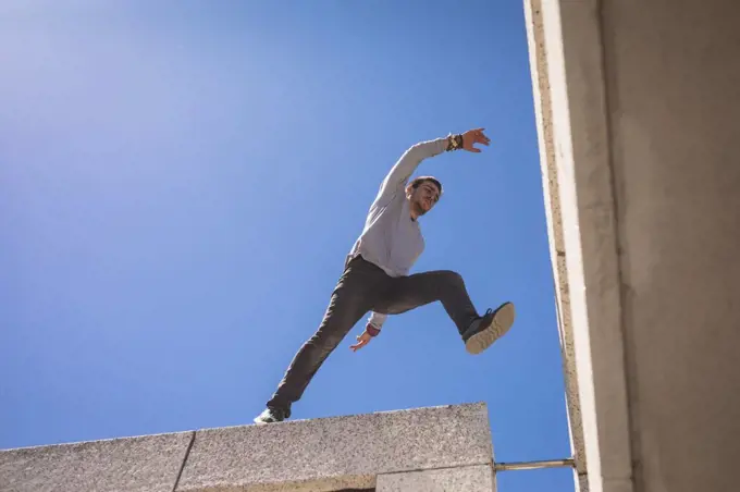 Front low angle view of a Caucasian man practicing parkour by the building in a city on a sunny day, jumping on the rooftop.