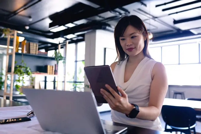 Front view of an Asian businesswoman wearing smart clothes, working in the modern office, standing by a desk and using her laptop computer and tablet. 