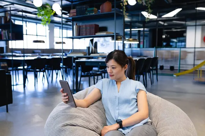 Front view of an Asian businesswoman wearing smart clothes, working in a modern office, sitting on a bean bag and using her tablet.