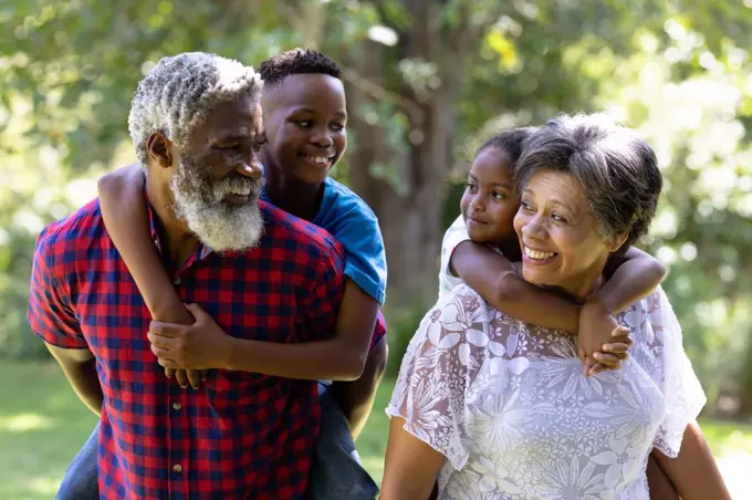 Senior mixed race couple enjoying their time at the garden, holding their grandchildren piggy back, looking at each other and smiling, on a sunny day