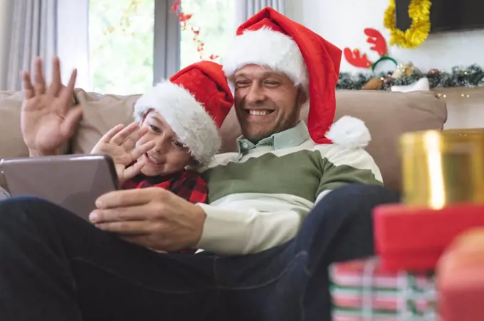 Caucasian man at home with his son at Christmas, wearing Santa hats sitting on sofa in living room, using digital tablet, waving. Social distancing during Covid 19 Coronavirus quarantine lockdown.