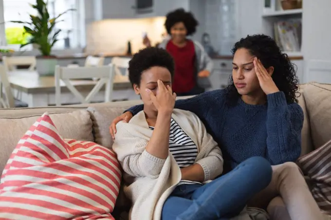 Upset mixed race lesbian sitting on couch. daughte rin the background. self isolation quality family time at home together during coronavirus covid 19 pandemic.