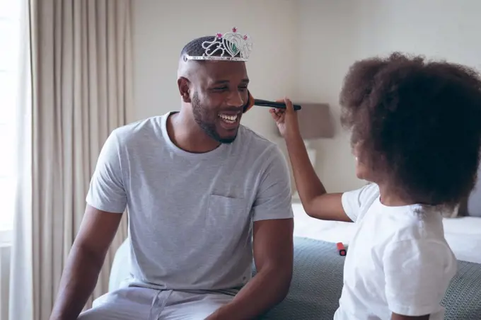 African american man wearing tiara having makeup put on by his daughter. staying at home in self isolation during quarantine lockdown.