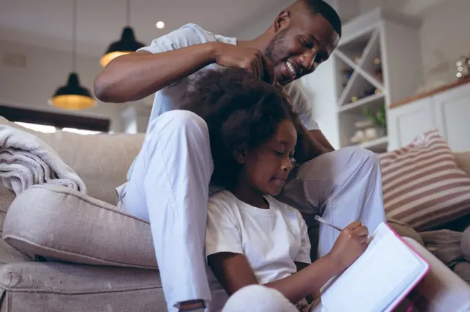 African american man sitting on bed doing his daughter hair. staying at home in self isolation during quarantine lockdown.