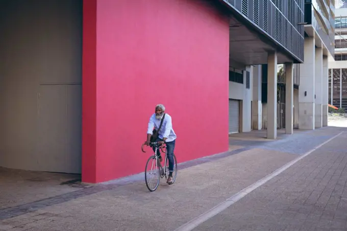 African american senior man riding bicycle in the street past red wall. digital nomad out and about in the city.