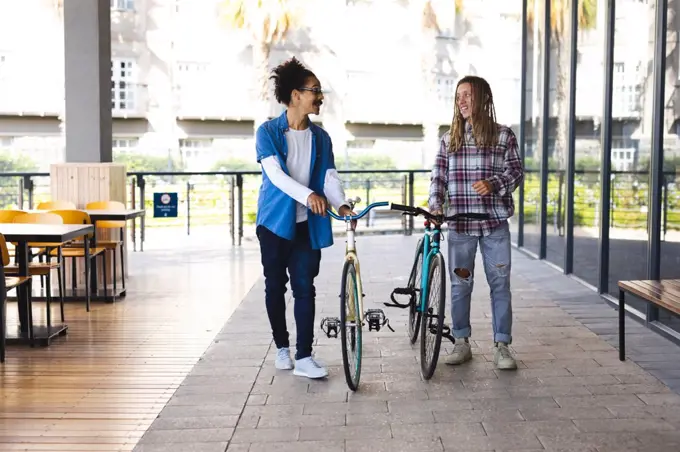 Two happy mixed race male friends wheeling bicycles in the street and talking. green urban lifestyle, out and about in the city.