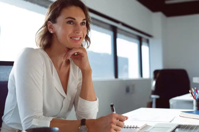 Smiling caucasian businesswoman sitting at desk, making notes at work. independent creative business at a modern office.