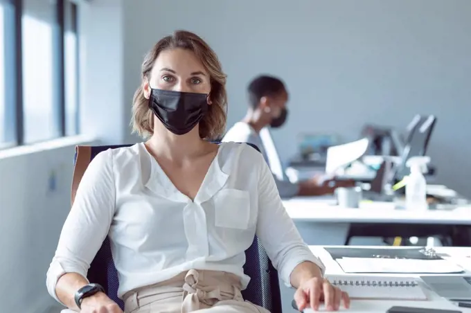 Caucasian businesswoman wearing face mask, sitting at desk, looking at camera. independent creative business at a modern office during coronavirus covid 19 pandemic.