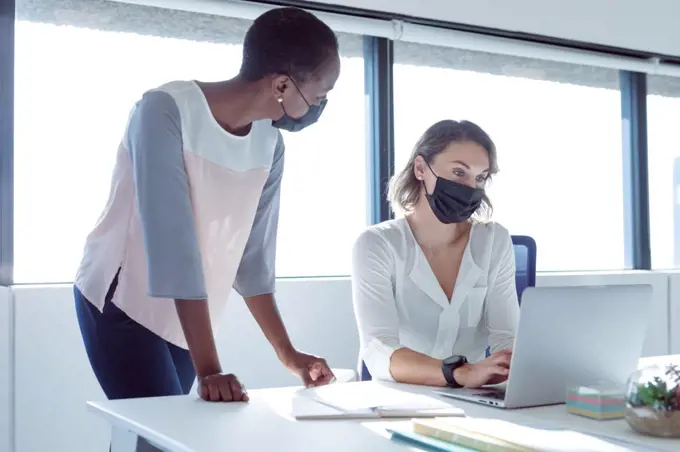 Two diverse smiling businesswomen wearing face masks, working together, using laptop, talking. independent creative business at a modern office during coronavirus covid 19 pandemic.