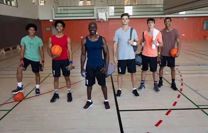 Portrait of diverse male basketball team and coach smiling and holding balls. basketball, sports training at an indoor court.