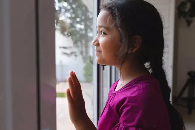 Happy hispanic girl standing at window with hand on glass, looking out and smiling. free time at home.