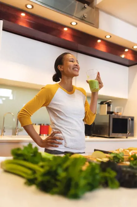 Smiling mixed race woman in kitchen drinking health drink. domestic lifestyle, enjoying leisure time at home.