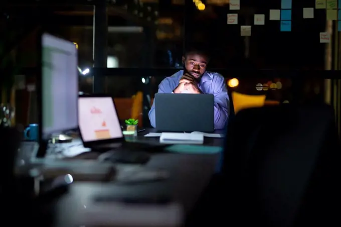 African american businessman working at night, sitting at desk and using laptop. working late in business at a modern office.