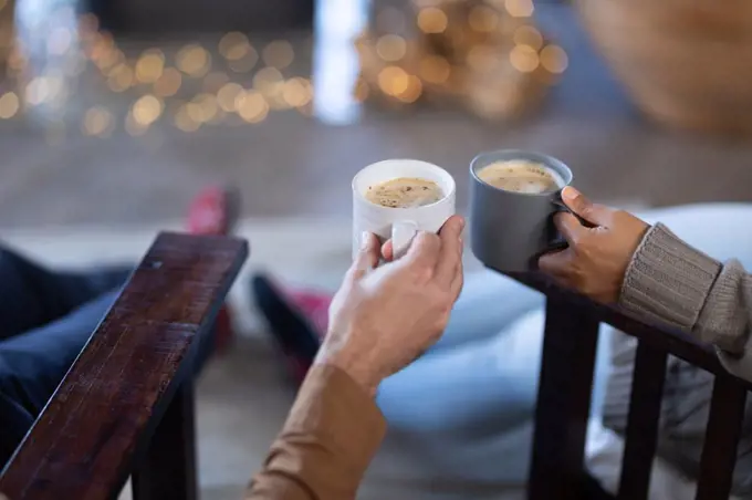 Happy diverse couple in living room holding mugs and drinking coffee. spending time off at home.