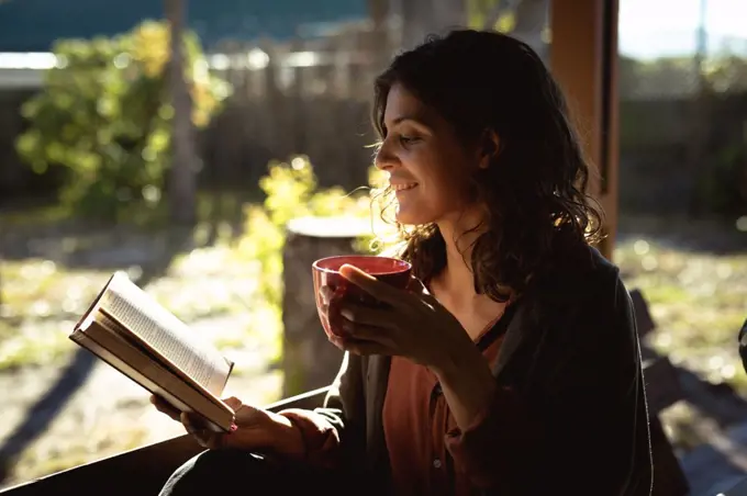 Mixed race woman reading book and drinking coffee in sunny garden. healthy lifestyle, enjoying leisure time at home.