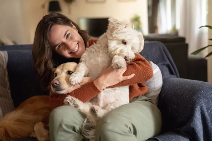 Portrait of smiling caucasian woman in living room sitting on sofa embracing her pet dogs. domestic lifestyle, enjoying leisure time at home.