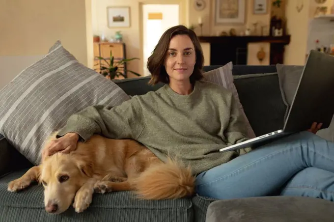 Portrait of smiling caucasian woman in living room, sitting on sofa with her pet dog, using laptop. domestic lifestyle, enjoying leisure time at home.