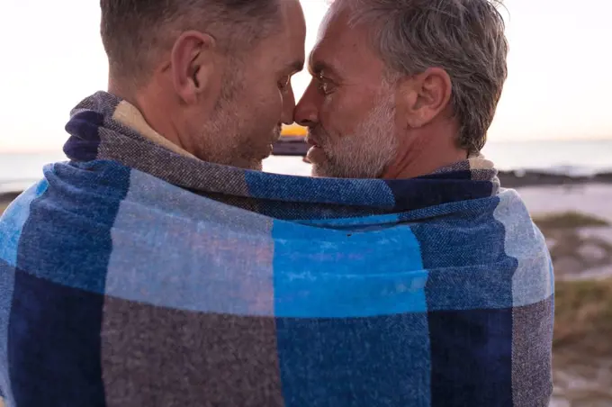 Happy caucasian gay male couple wrapped in blanket, embracing and taking selfie on beach at sunset. summer road trip and holiday in nature.