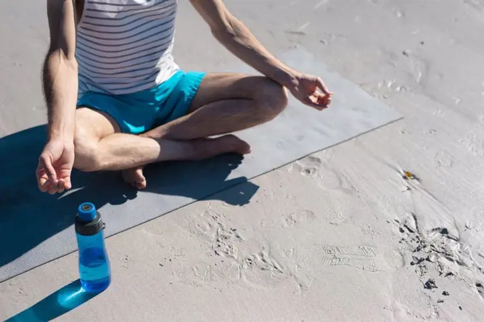 Low section of man meditating while practicing yoga by water bottle on sand at beach. fitness and healthy lifestyle.