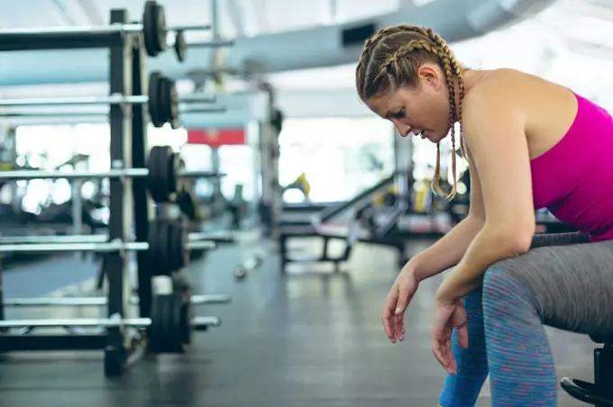 Side view of thoughtful young Caucasian female athletic relaxing on a bench in fitness center. Bright modern gym with fit healthy people working out and training