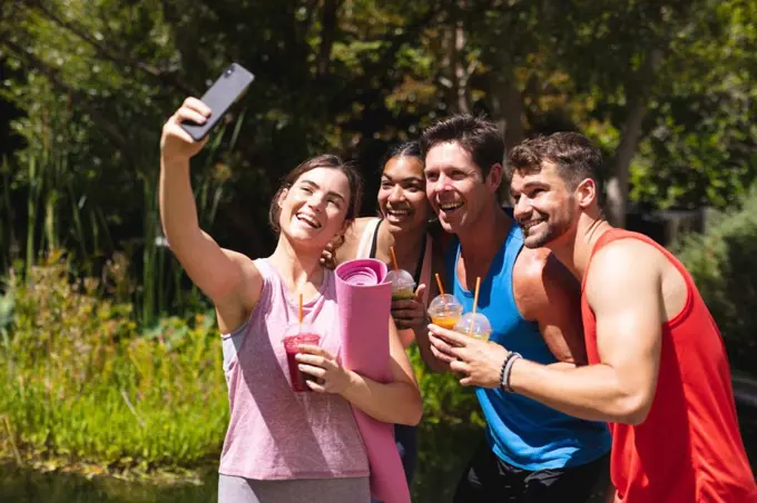 Happy male and female friends taking selfie while holding healthy drinks post workout in park. healthy lifestyle, drink, wireless technology and fitness.