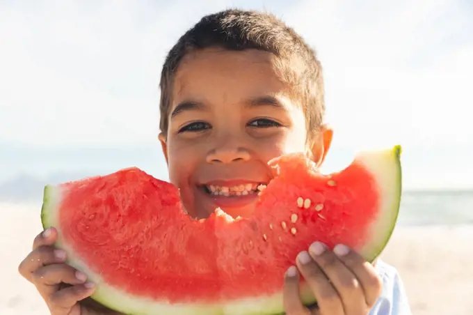 Portrait of smiling cute biracial boy holding large fresh watermelon slice at beach on sunny day. childhood and healthy food.