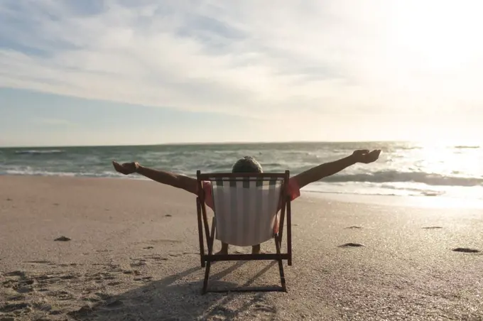 Retired senior biracial man with arms outstretched sitting on folding chair at beach during sunset. lifestyle and weekend.