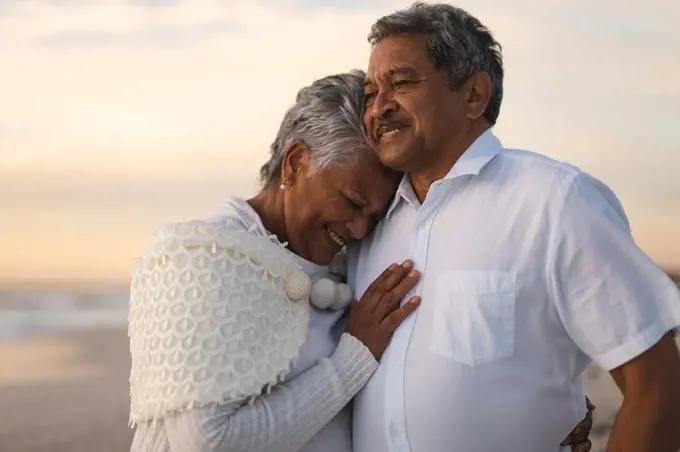 Smiling senior biracial woman embracing man looking away while standing at beach during sunset. lifestyle, love and weekend.