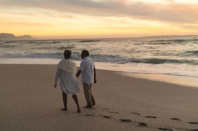 Full length of senior biracial couple walking together on shore at beach during sunset. lifestyle, love and weekend.