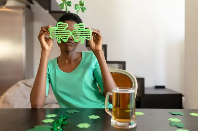 African american woman holding shamrock novelty glasses while sitting with beer mug at table. unaltered, people, emotions, lifestyle and st patrick's day celebration concept.