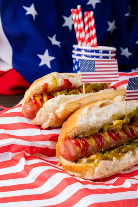 Close-up hot dogs with sauces and jalapenos and american flag, straw and disposable cups on table. unaltered, unhealthy food, meat, bread, sausage and fast food.