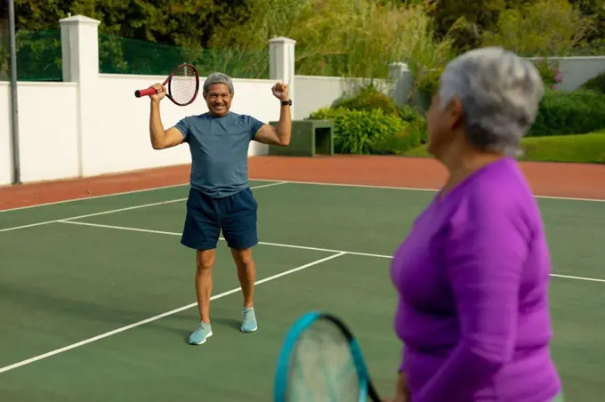 Biracial senior woman looking at cheerful senior man holding racket and gesturing in tennis court. unaltered, sport, competition, togetherness, achievement, retirement, healthy lifestyle concept.