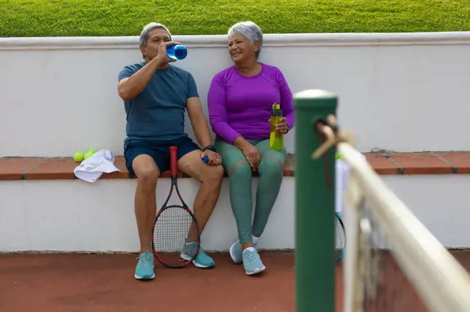 Full length of tired biracial senior couple drinking water while sitting on bench at tennis court. copy space, relaxation, unaltered, sport, togetherness, love, retirement, active lifestyle.