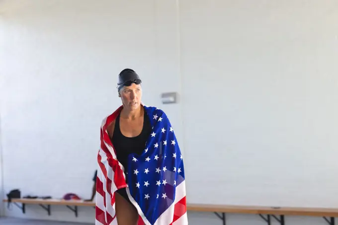 Caucasian female athlete swimmer wrapped in an American flag, with copy space. She stands confidently in a gym setting, suggesting athletic pride or patriotism.