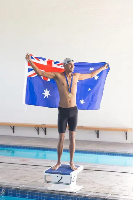 Young biracial male swimmer celebrates victory at a swimming pool with the Australian flag. Holding the Australian flag, the athlete stands proud with a medal, symbolizing triumph.