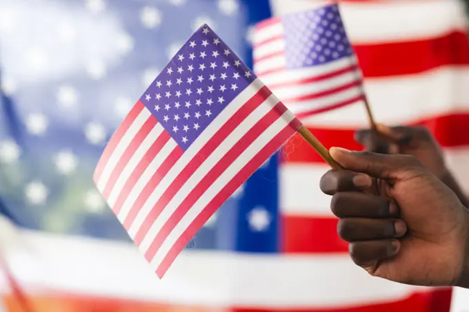 African American hand holding an American flag with another flag in the background. The focus on the flag symbolizes patriotism and national pride.