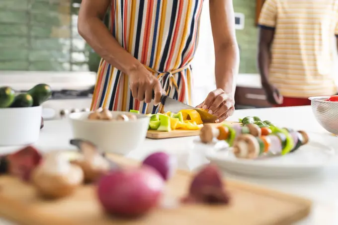 Biracial woman in a striped dress is chopping vegetables in a kitchen. Fresh produce and a focus on healthy eating set the scene for meal preparation.