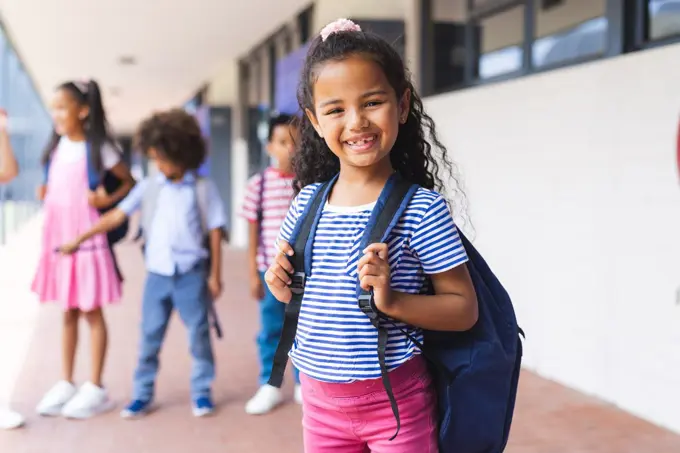 Students, including biracial girl and African American boy, smiling with backpacks. Standing outside school building, ready for a day of learning and fun, unaltered