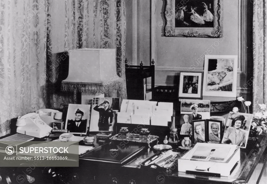 Clarence House - Princess Elizabeth's London Home -- Princess Elizabeth's writing desk in her Sitting Room at Clarence House, the London residence of the Princess and the Duke of Edinburgh. The desk is of the Chippendale period. The photographs are of the Queen, Queen Mary, Princess Margaret and several of the Duke of Edinburgh, including one taken during the war when he wore a beard. Beyond are photographs taken at the christening of Prince Charles, and a painting of the ballet Le Iac des Cygnes, by Oliver Messel. October 28, 1949.