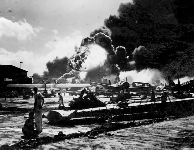 Against a background of smoke and flame, wreckage-strewn Naval Air Station is shown after one of the sneak attacks by Jap bomber on Pearl Harbor, on December 7th, 1941. Note wing of blasted patrol bomber, (foreground) huge ball of flame (background) came from explosion that happened just as this photo was taken. Picture was just released by the Navy Department in Washington. February 4, 1943.